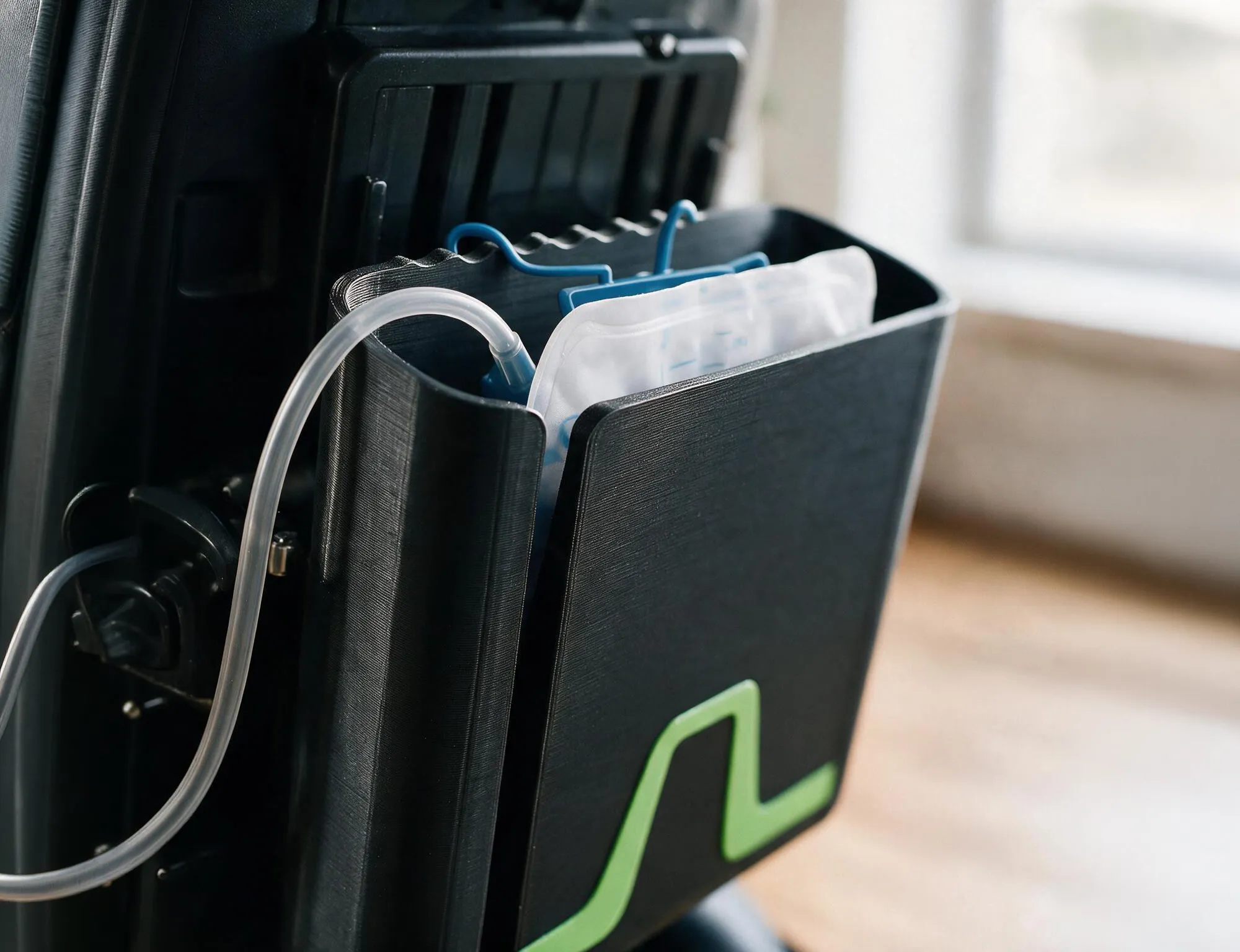 Nahaufnahme des Flowcase am Rollstuhl: Der Katheterbeutel hängt mit seinen blauen Haken an der gewellten Innenkante, die glatte Außenwand verdeckt ihn fast vollständig.
