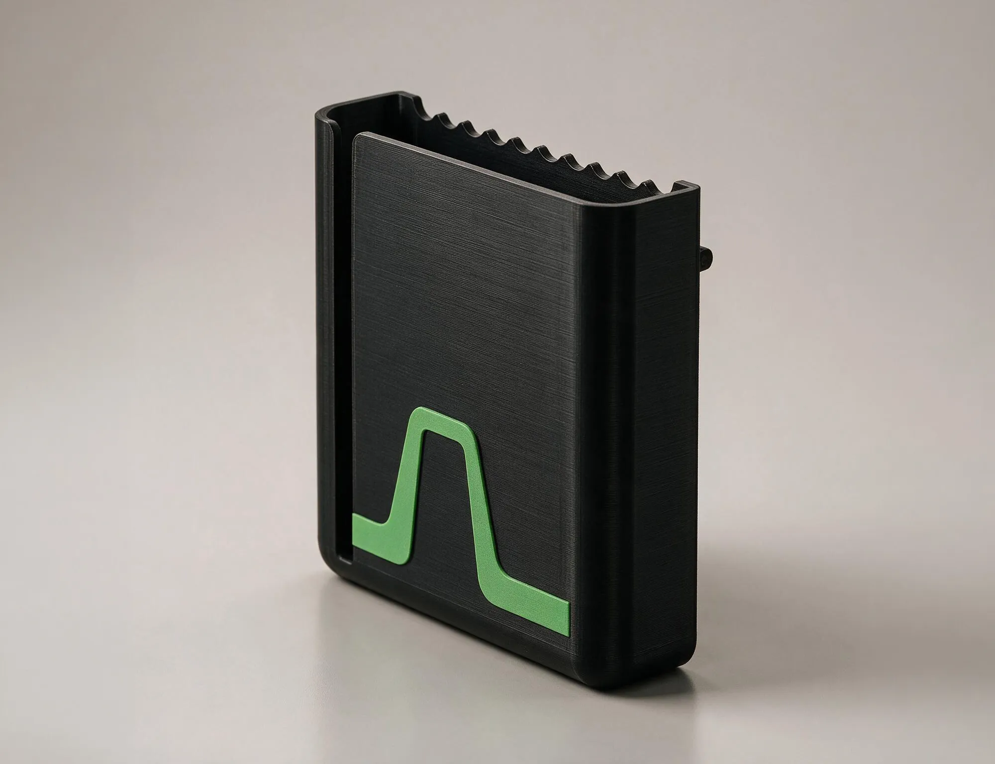 Der Flowcase freistehend vor grauem Hintergrund: mattschwarzes 3D-gedrucktes Gehäuse mit sichtbaren Druckschichten, gewellter Einhängekante innen und grüner Akzentlinie auf der Außenwand.