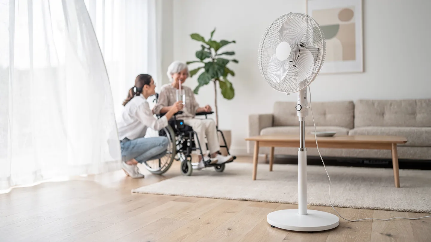 Ein weißer Standventilator steht in einem hellen Wohnzimmer. Im unscharfen Hintergrund kniet eine junge Frau neben einer älteren Frau im Rollstuhl, die aus einem Glas mit Trinkhalm trinkt.